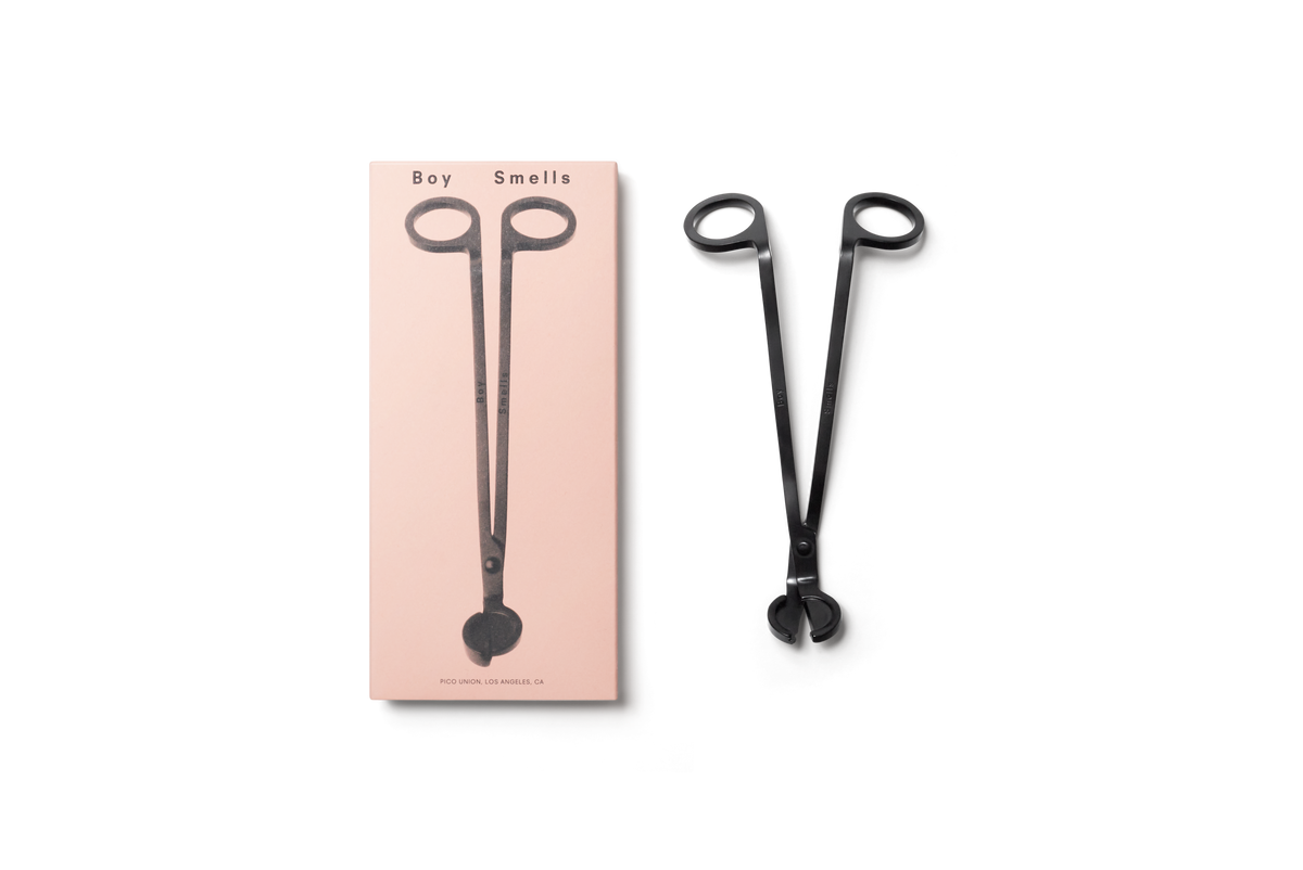 product shot of exterior pink box with black text, boy smells logo in black and black image of wick trimmer , next to box is metallic black wick trimmer with engraved boy smells logo against a neutral background. 