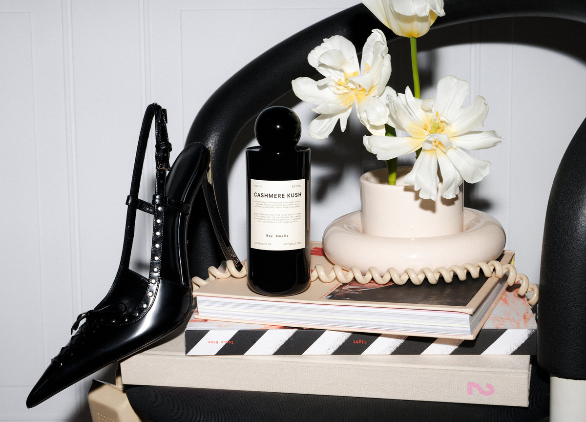Editorial image of Cashmere Kush Room spray sitting on a stack of books on a chair surrounded by a modern white vase and black high heel and a phone cord hanging off edge.